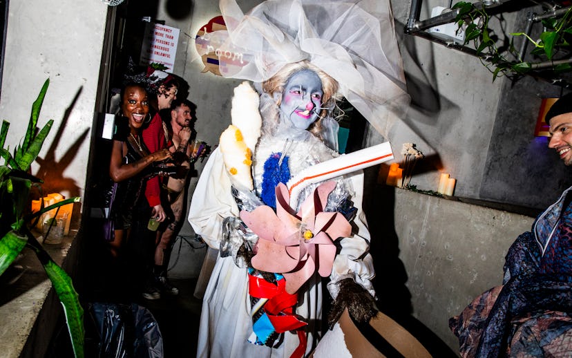 A person dressed in elaborate make-up and a large hat carries colorful decorations, while a group of...