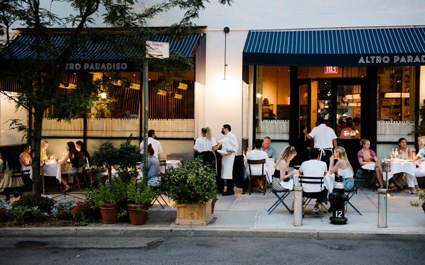 People dining outside a restaurant with a blue awning, named "Altro Paradiso," on a tree-lined urban...