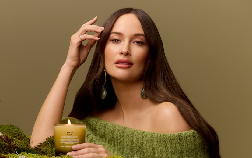 A woman in a green off-shoulder top poses with a lit candle on a bed of moss, exuding a serene ambia...