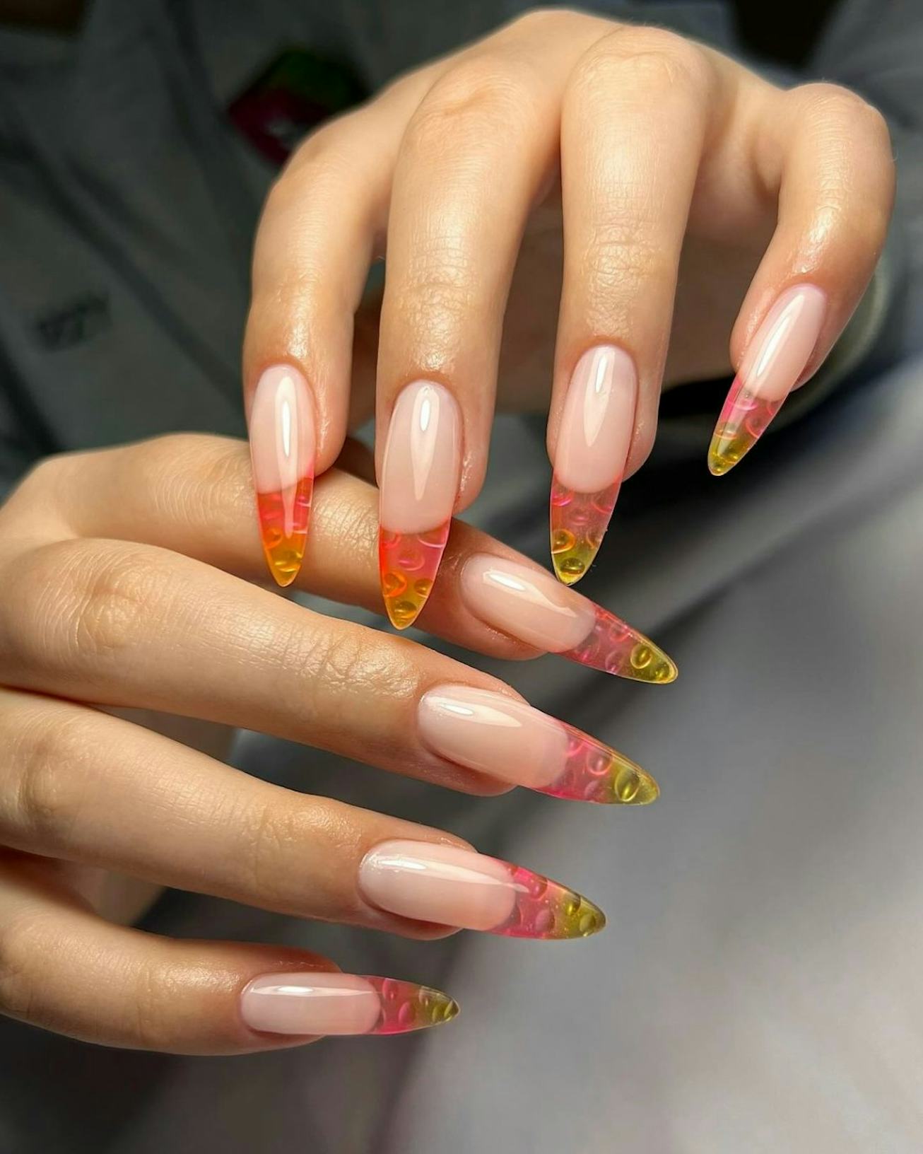 A close-up of hands showcasing long, almond-shaped nails with a glossy nude base and vibrant orange ...
