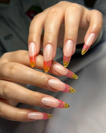 A close-up of hands showcasing long, almond-shaped nails with a glossy nude base and vibrant orange ...