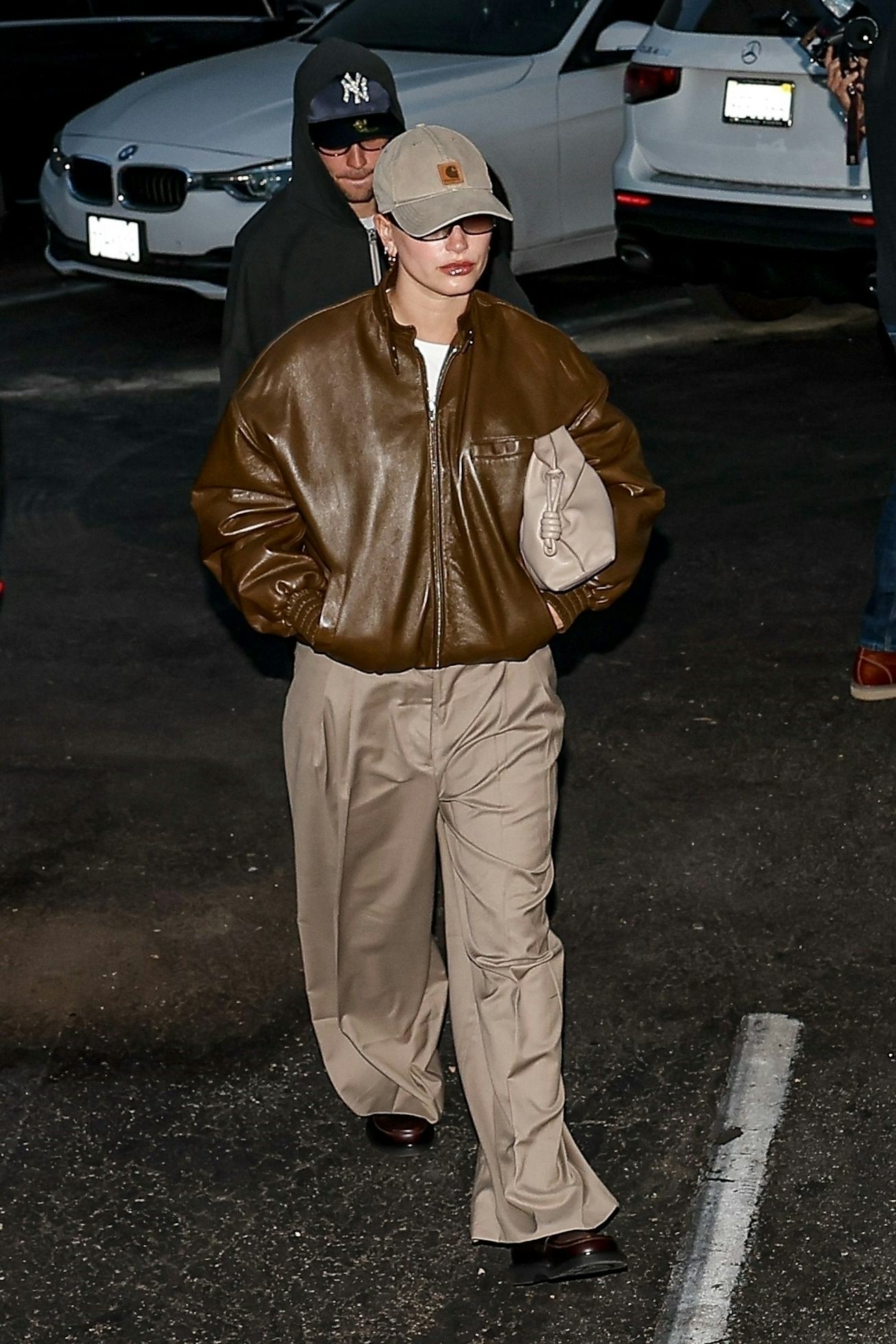 A person wearing a brown leather jacket and beige pants walks confidently in a parking lot, sporting...