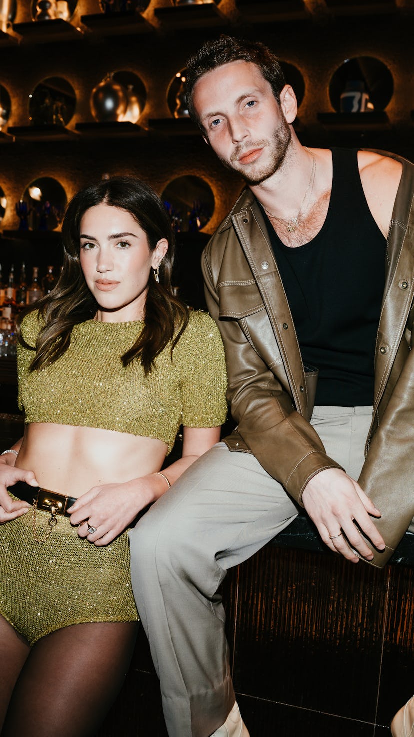 A woman in a sparkling green outfit and a man in a sleeveless shirt sit at a bar, surrounded by illu...