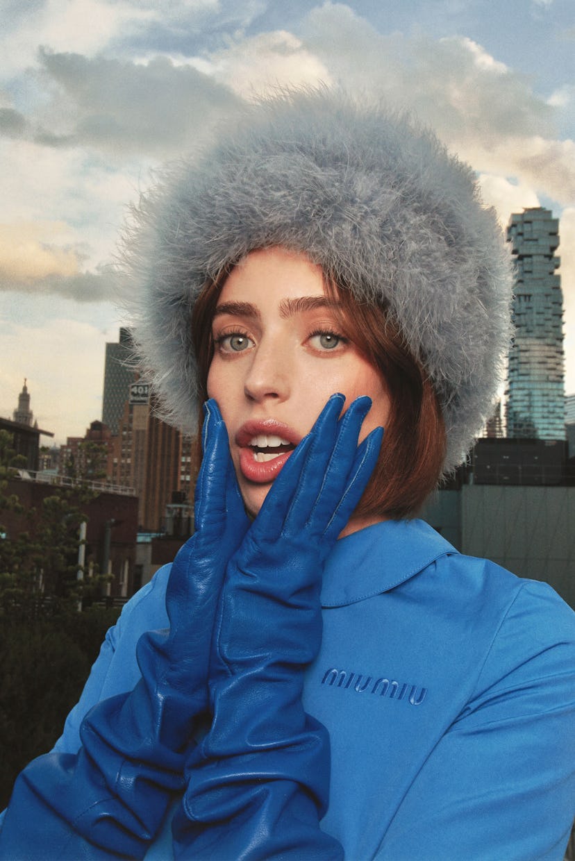 A woman in a fur hat and vibrant blue gloves poses with a surprised expression against a city skylin...