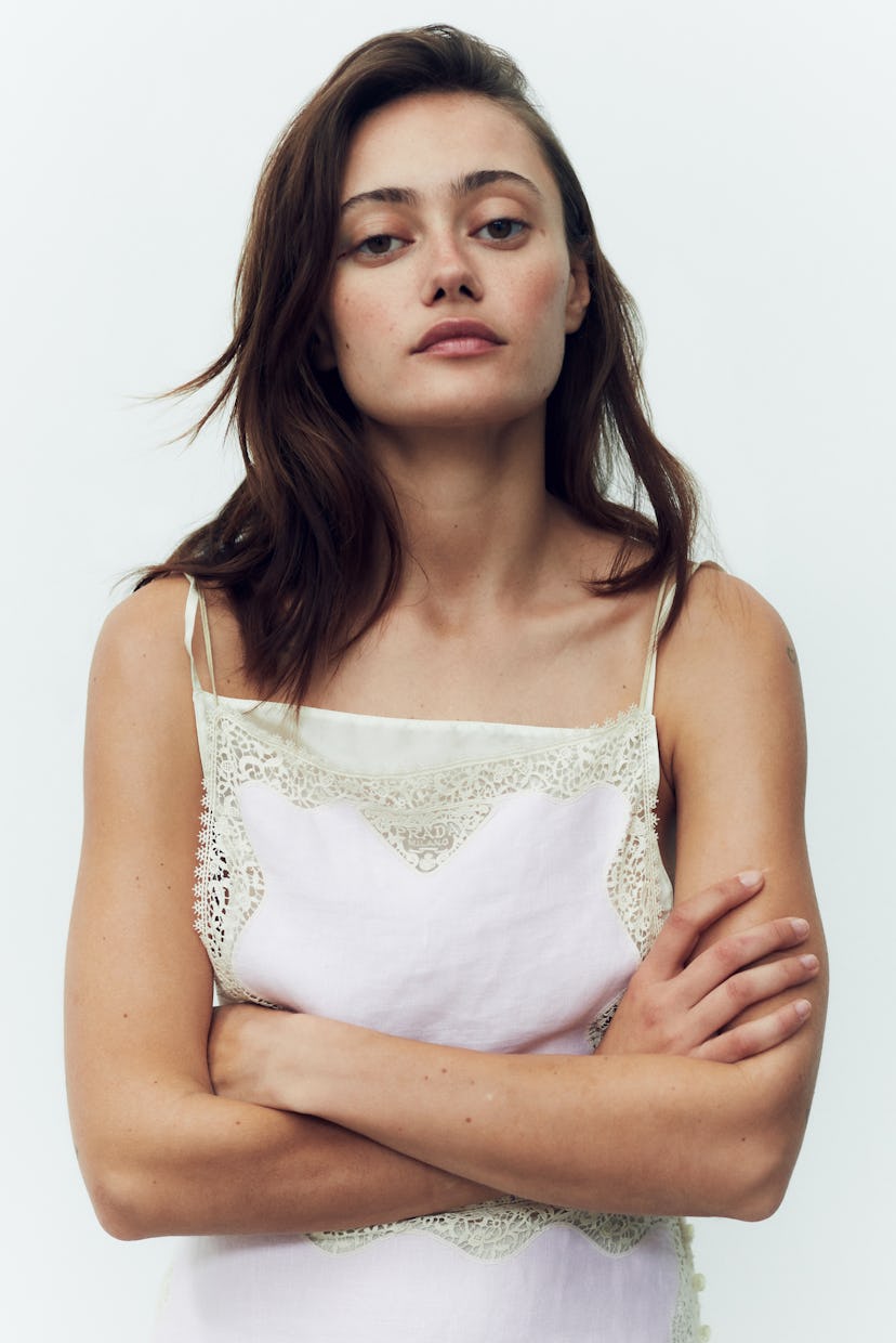 Portrait of a woman with arms crossed, wearing a white lace-trimmed top, against a plain background.