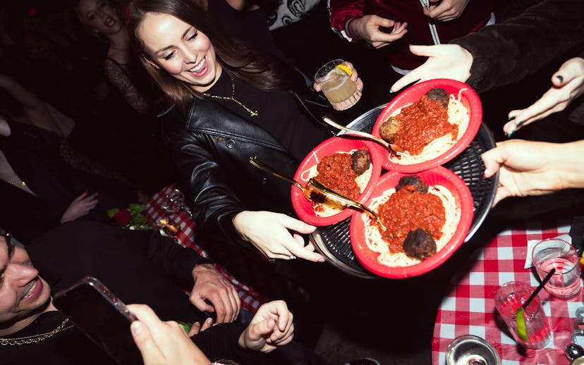 People enjoying a party, sharing plates of spaghetti and meatballs, with one person smiling and hold...