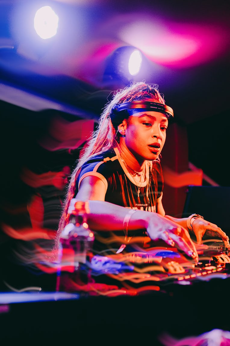 A focused DJ mixes tracks on stage under colorful lights, wearing a headband and earrings.