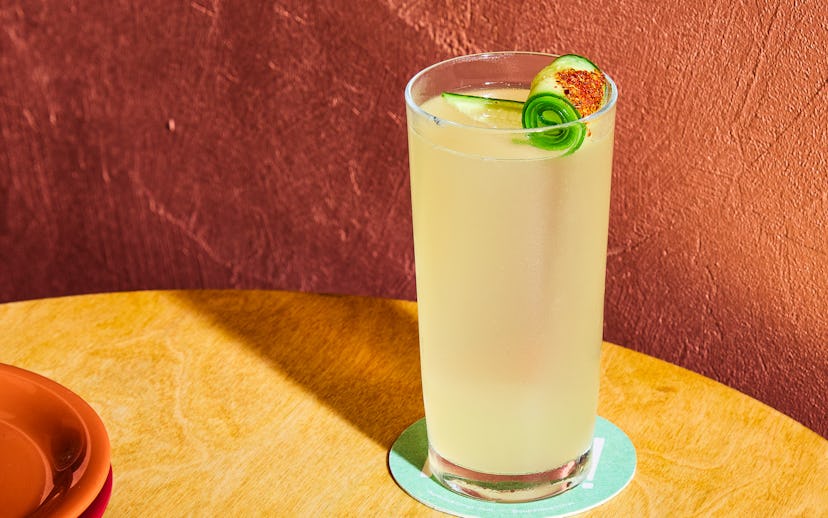 A glass of lemonade with a lime slice and cucumber on a sunny table, with a red textured wall behind...