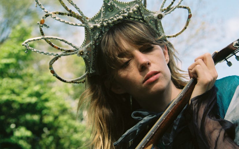 A person with long, light brown hair, wearing an ornate crown, thoughtfully playing a violin outdoor...