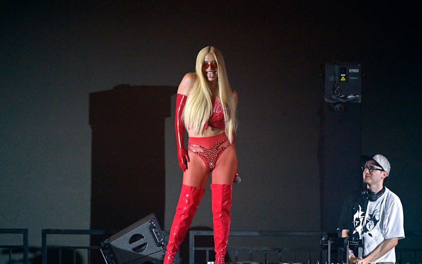A performer in a red outfit with long blonde hair and red boots on stage, with a male DJ in the back...