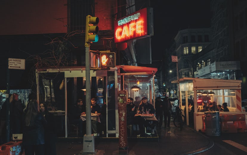 Night scene of a busy street corner with people dining at outdoor cafe booths, under a neon "Cafe" s...