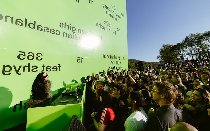A large crowd gathers in front of a vibrant green wall with text. A DJ performs on stage, energized ...