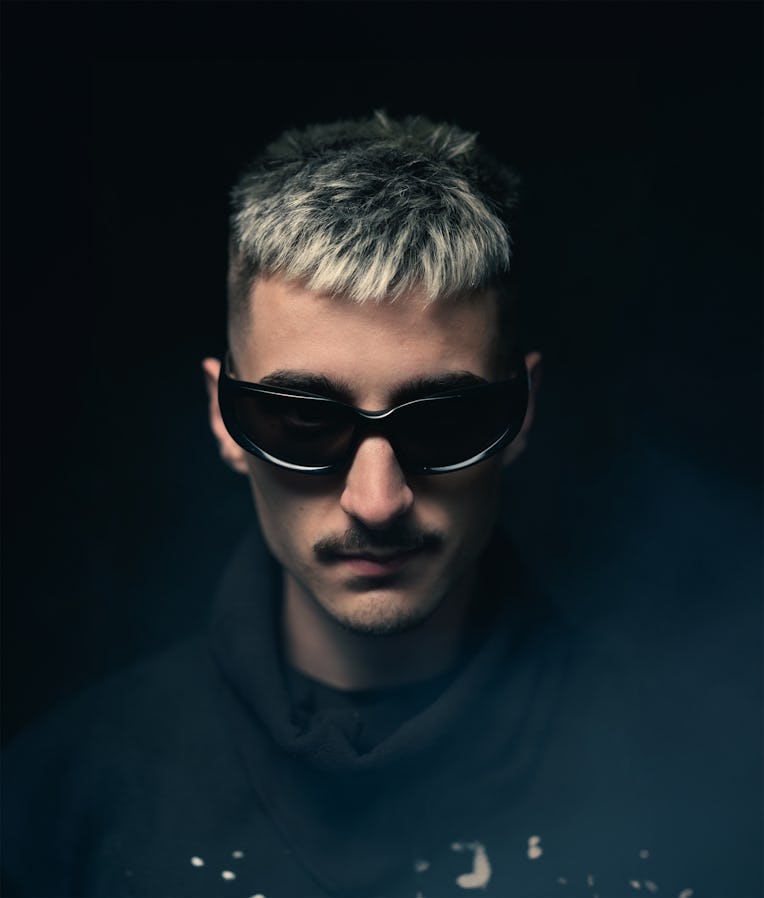 A close-up portrait of a person with short, styled light grey hair, wearing dark sunglasses and a bl...