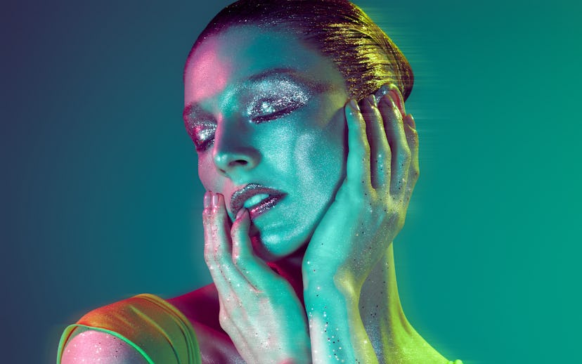 A close-up portrait of a person with shimmering skin and glittery eye makeup, illuminated by vivid g...