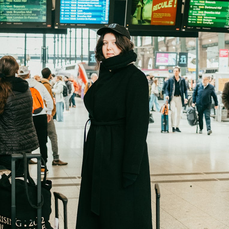 A young woman in a black coat and cap stands at a busy train station, surrounded by travelers and di...
