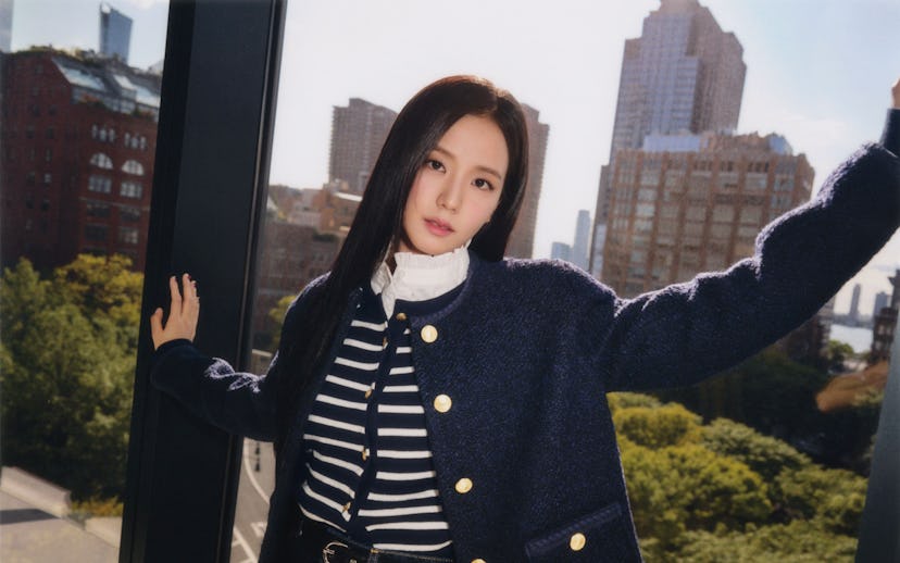 A young woman poses elegantly by a large window, wearing a navy jacket over a striped top, with city...